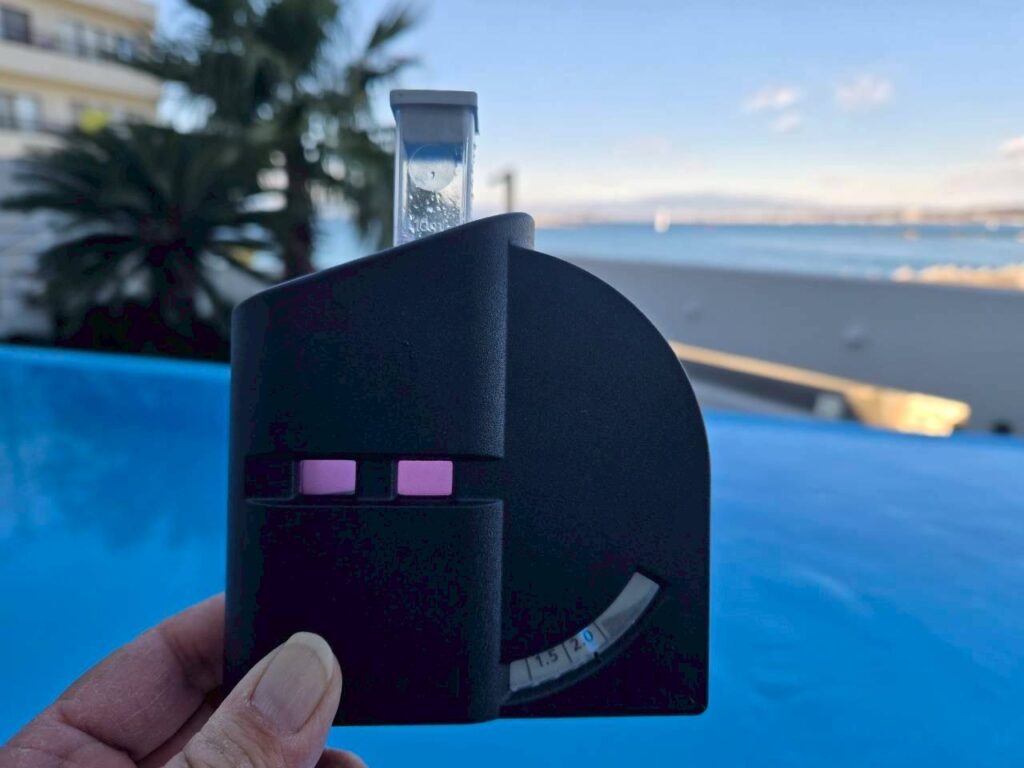 手に持った水質測定器の透明チューブに採取した水が入り、背後にはプールとヤシの木が並ぶ屋外空間が広がっている。