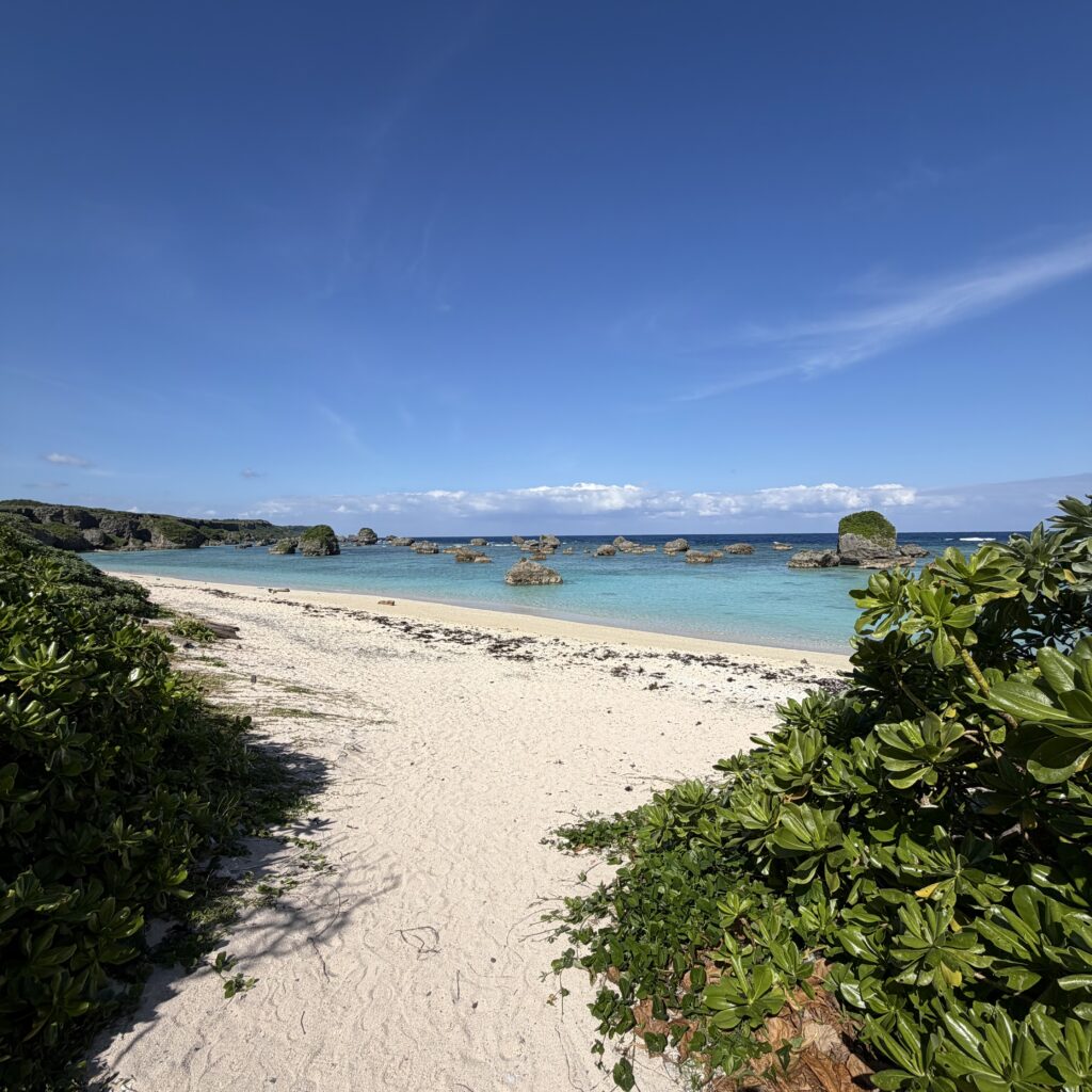 白砂の浜辺と透明なエメラルド色の海、岩場が点在する宮古島のような南国のビーチ風景。青空と海岸線が広がる自然の景観