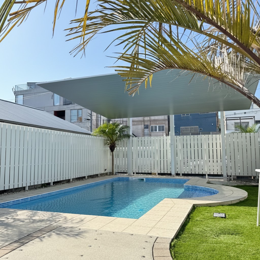 白い縦格子フェンスに囲まれた都市型の屋外プール。人工芝とタイルデッキが整備され、ヤシの木とカーポートSCがリゾート感を演出する。